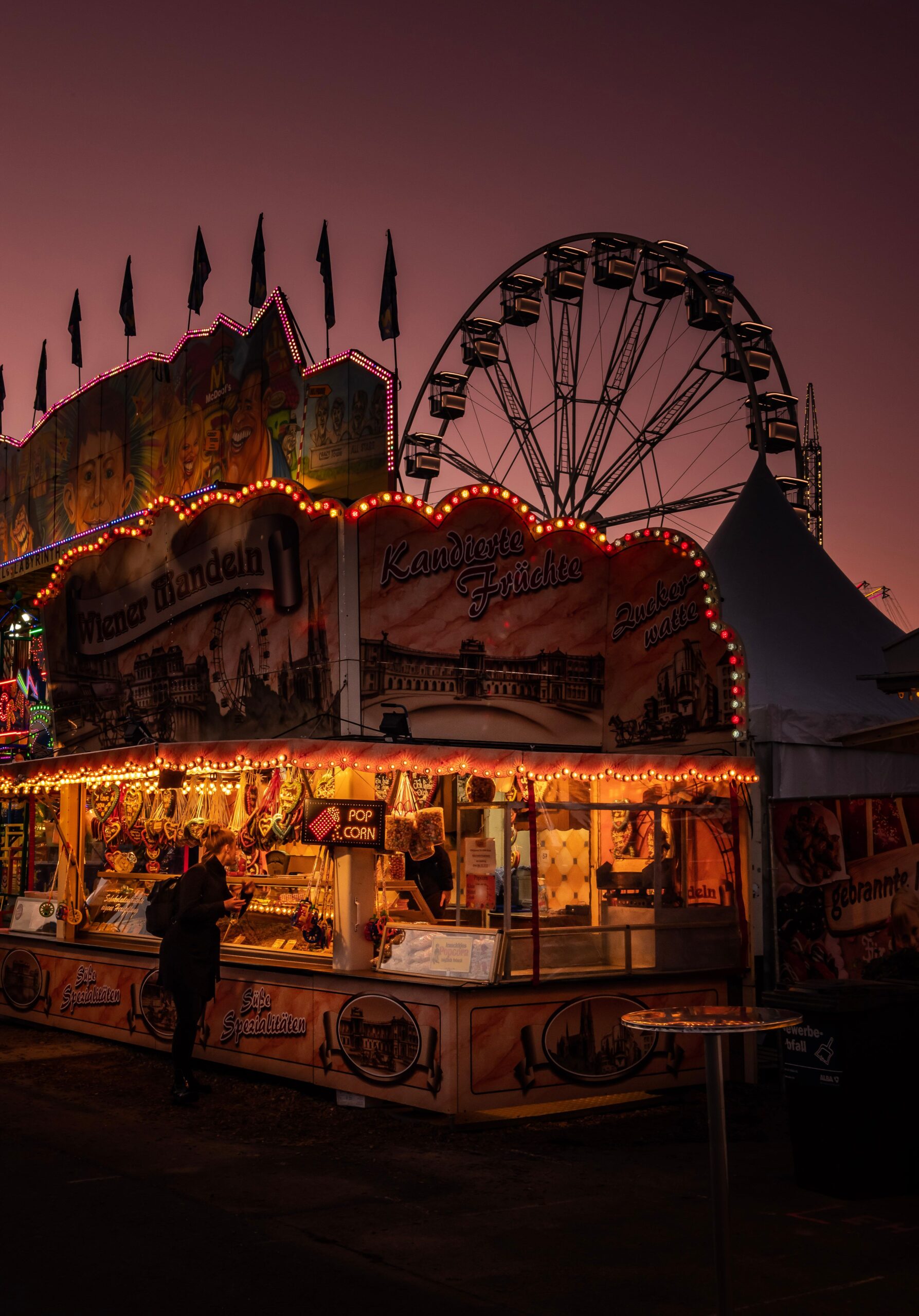 A carnival stand is lit up in front of a Ferris wheel and a sunset.
