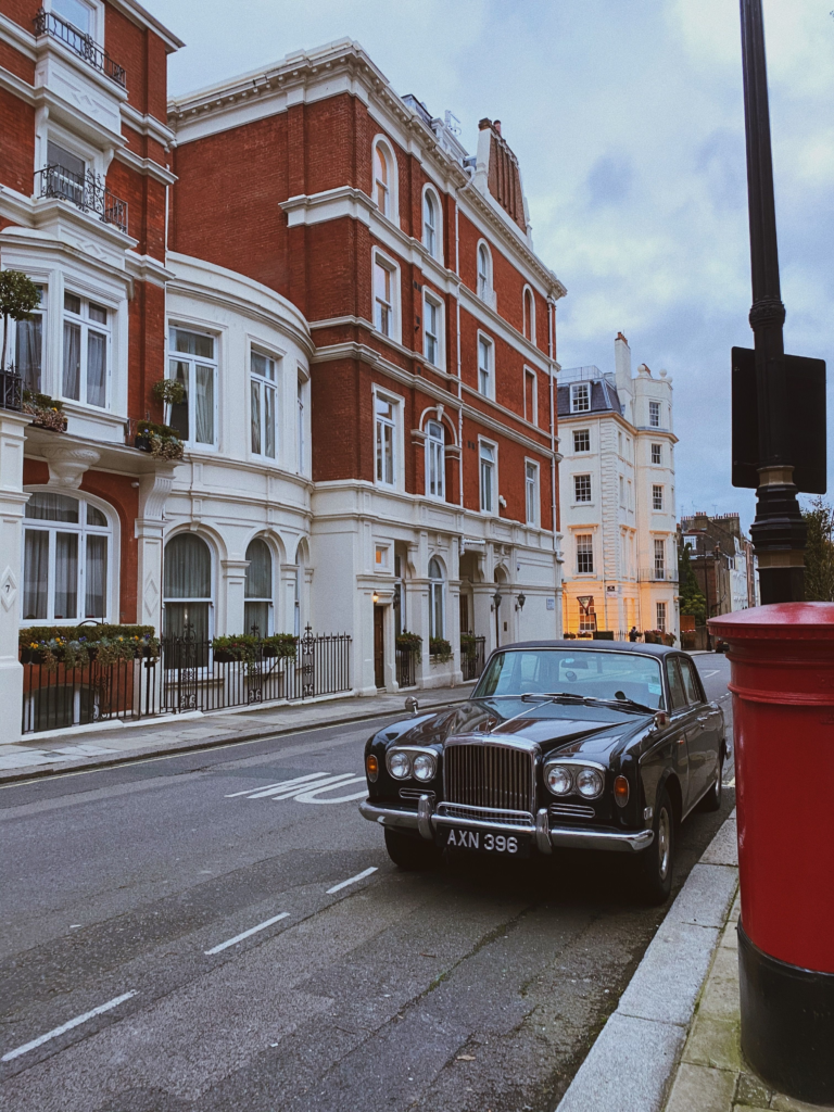 An old car sits on a road in London next to brick buildings.
